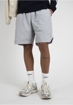 Mies pukeutunut vaaleanharmaisiin shortseihin, valkoisiin merkkisukkiin ja hopeisiin juoksukenkään, seisoo yhdellä kädellä taskussa yksiväristä taustaa vasten.