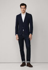 Marineblazer mit Revers, zwei Knöpfen, kombiniert mit einem weißen Poloshirt, dunklen Jeans und braunen Lederschuhen. Glatte Textur, taillierte Passform.