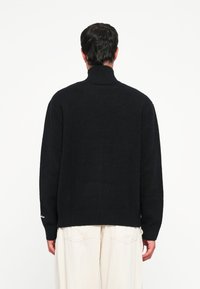 Suéter negro de cuello alto con una textura suave, puños y dobladillo acanalados, y una silueta suelta. Mostrado por la parte de atrás, combinado con pantalones claros.