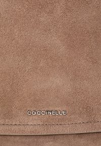 Borsa a tracolla in suede marrone con una texture liscia, dotata di un logo metallico "Coccinelle" e dettagli di cucitura sottili.