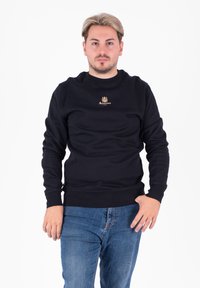 Czarna bluza z okrągłym dekoltem, z ribbowanymi mankietami i dołem. Z�łote haftowane logo na piersi. Noszona z niebieskimi dżinsami.