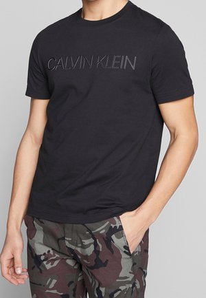 Camiseta de algodón negra con mangas cortas, que presenta un logo "CALVIN KLEIN" brillante y en relieve en el pecho; combinada con pantalones de camuflaje.