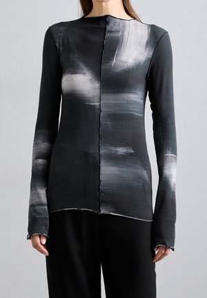 Žena nosící přiléhavé černé tričko s dlouhým rukávem, se šedobílými abstraktními štětcovými vzory a vystaveným středovým švem.