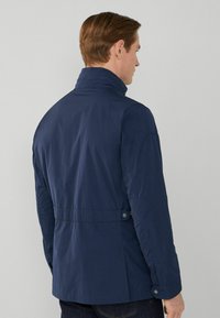 Mörkblå lättviktig jacka med ståkrage, tryckknappar, justerbar midja och slät textur. Inkluderar sidofickor och en skräddarsydd passform.
