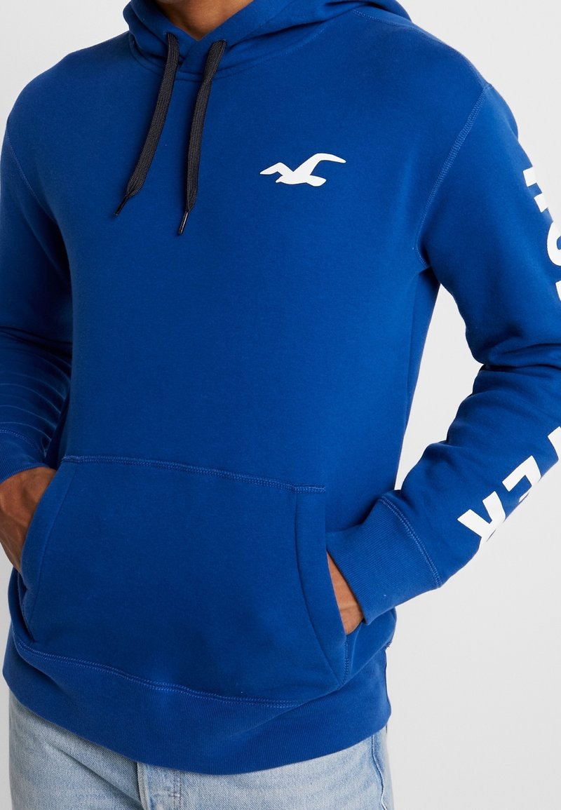 Blue hoodie made of soft fabric, featuring a black drawstring, white logo on chest, and text on the sleeve. Kangaroo pocket at the front.