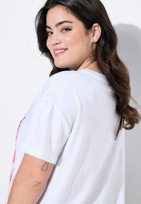 Camiseta de algodón blanca con mangas cortas que presenta un diseño gráfico colorido en la parte frontal. El modelo tiene el cabello largo y oscuro y lleva un pendiente de aro.