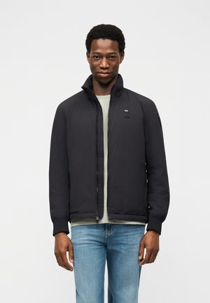 Bărbat purtând o jachetă neagră cu fermoar peste un tricou gri deschis și blugi albastru deschis, stând în fața unui fundal alb simplu.