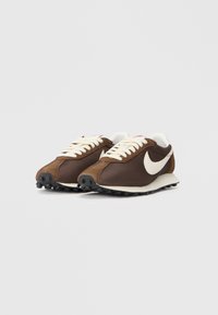 Brūni sporta apavi ar sieta un zamšāda augšdaļu, krēmkrāsas šņores, baltu Nike zīmi un strukturētu melnu gumijas zoli.