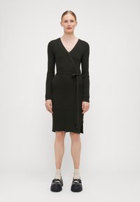 Mørkegrønn ribbestrikket kjole med V-hals, lange ermer, omslagsdesign, knytbelte og sideslits. Bæres med beige sokker og kraftige sorte sko.