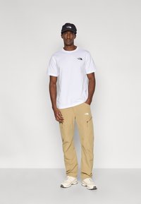 Άσπρο βαμβακερό μπλουζάκι με λογότυπο, μπεζ παντελόνι cargo με τσέπες με φερμουάρ και μαύρο καπέλο. Παπούτσια είναι αθλητικά ελαφρού χρώματος.