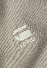 Tela de algodón beige con un logo bordado en blanco que dice "G-Star RAW", con una textura suave y un ligero relieve.