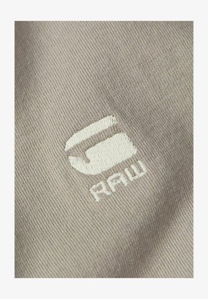 Tessuto di cotone beige con un logo ricamato in bianco che recita "G-Star RAW", con una texture morbida e una leggera rigatura.