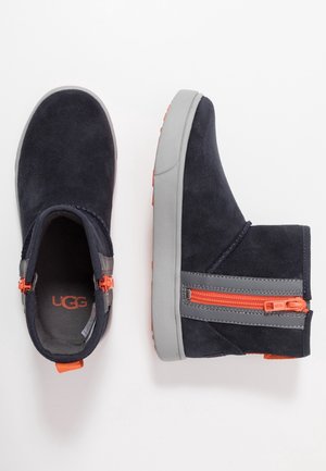 Paire de bottes en daim noir avec semelles grises et fermetures éclair latérales orange vif, une botte vue de dessus, l'autre de côté.