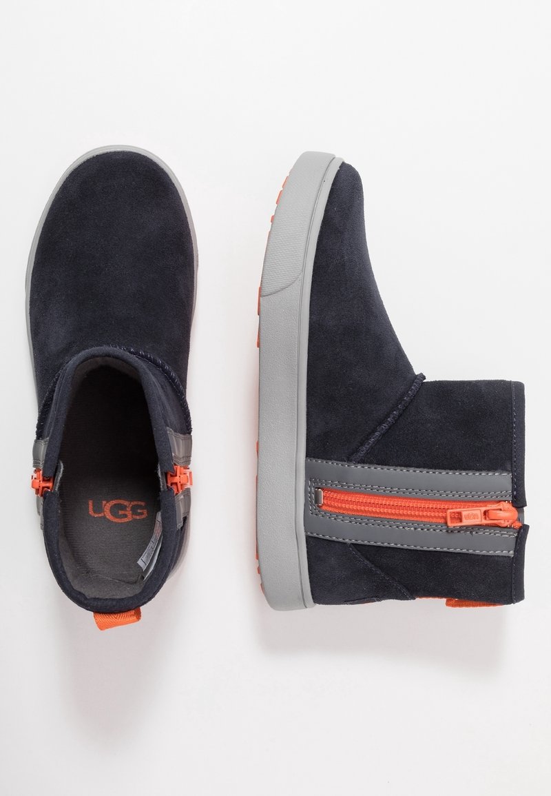 Paire de bottes en daim noir avec semelles grises et fermetures éclair latérales orange vif, une botte vue de dessus, l'autre de côté.