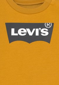 Mosterdgele sweatshirt met een geribbelde ronde hals, voorzien van een grijs logo met witte tekst, gecentreerd op de voorkant.