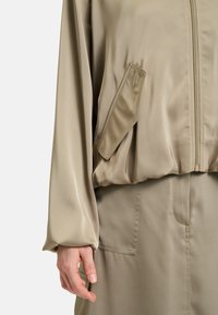 Giacca bomber in satin beige con maniche lunghe e chiusura a zip. Presenta una tasca a pattina sul lato sinistro e una vestibilità comoda. Texture liscia.