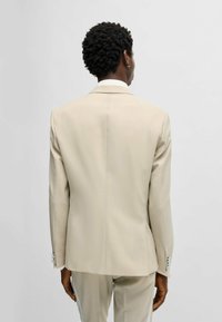 Giacca da abito di un beige chiaro con una vestibilità tailored, apertura singola e un leggero luccichio; presenta una superficie liscia e un design delle spalle strutturato.