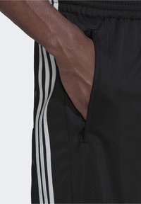 Hand in pocket of black sports pants with white side stripes and snap buttons, showing zipper on pocket.