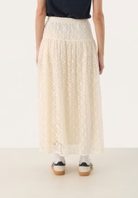 Jupe longue en dentelle crème avec un motif en losanges, taille élastique et design superposé, associée à des chaussures décontractées et des chaussettes blanches.