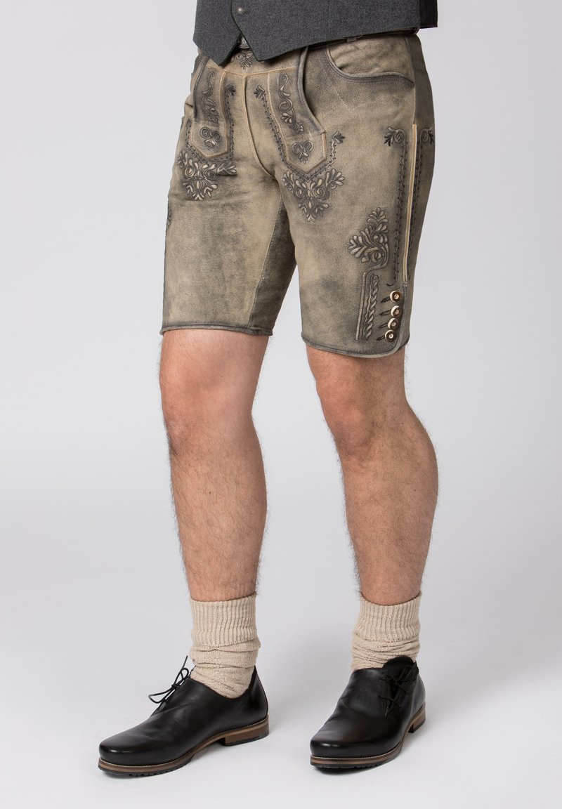 Pantaloni lederhosen in pelle marrone chiaro con cuciture decorative, tasche frontali e dettagli in bottone lungo il lato, abbinati a scarpe nere e calzini beige.