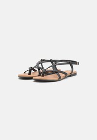 Black flat sandals with cross-strap design, featuring a tan sole and ankle strap with buckle detail. Smooth texture, lightweight construction.