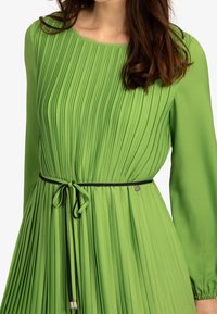 Vestido verde con plisados, de mangas largas y escote redondo. Presenta un cinturón negro con lazo en la cintura y una textura suave y fluida.