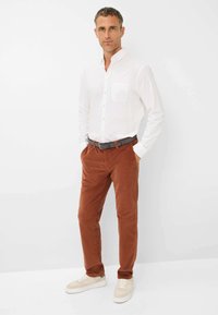 Chemise blanche à boutons, pantalon en velours côtelé marron, ceinture grise et chaussures légères. Textures lisses et coupe ajustée.