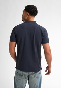 Námořnická bavlněná polo košile s krátkým rukávem, límečkem a detailním logem na rameni. Nesená s světle modrými džíny.