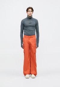 Jauns cilvēks, tērpies pelēkā garās piedurknes zip-up kreklā ar pirkstu caurumiem, oranžās plašās biksēs ar kabatām un baltās sporta apavos uz vienkārša fona.