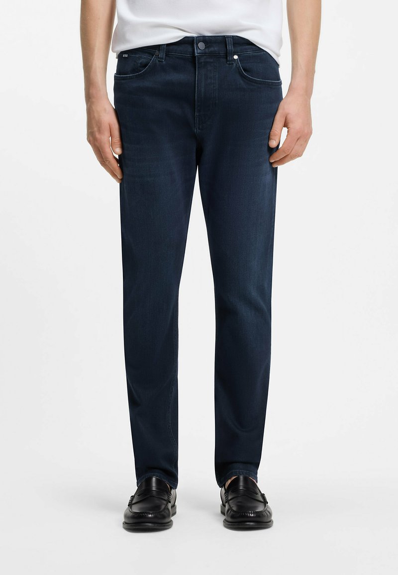 Homme portant un jean droit bleu foncé et des mocassins en cuir noir, debout devant un fond blanc uni.