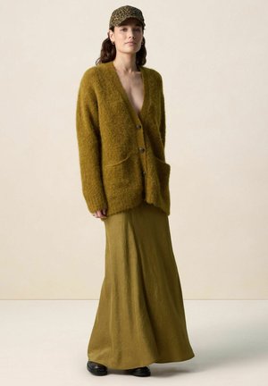 Femme portant un cardigan marron duveteux par-dessus une longue jupe assortie, des chaussures noires et une casquette à imprimé léopard, debout devant un fond uni.