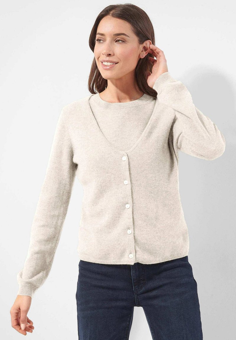 Frau lächelt, trägt eine cremefarbene, geknöpfte Strickjacke über einem passenden Oberteil und dunklen Jeans, mit einer Hand, die ihr Haar berührt, vor einem schlichten Hintergrund.