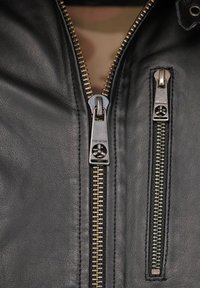 Schwarze Lederjacke mit zwei vorderen Reißverschlüssen, einem silbernen und einem goldenen. Glatte Textur mit sichtbaren Nähten um die Reißverschlüsse.