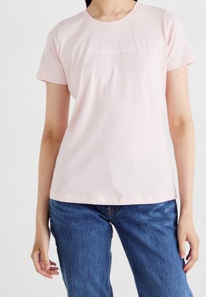 Personne portant un t-shirt rose clair à manches courtes avec un texte subtil et un jean bleu en denim, debout devant un fond blanc uni.
