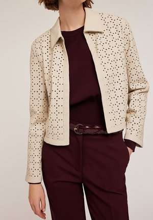 Kvinde iført beige jakke med sorte trekantede udskæringer over en mørk burgunderrød sweater og matchende bukser med bælte, stående med den ene hånd i lommen.