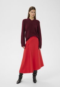 Bordeauxroter V-Ausschnitt-Pullover in lockerer Passform, kombiniert mit einem fließenden roten Maxirock mit diagonaler Drapierung, getragen mit schwarzen kniehohen Stiefeln.