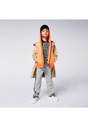 Detský outfit s béžovým kabátom, oranžovou kapucňou, čiernym grafickým tričkom, svetlošedými džínsy a bielymi teniskami.