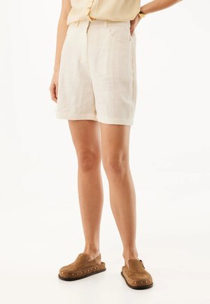 Žena v béžových vysokým pasem šortkách, světlejším tílku bez rukávů, hnědých nazouvacích botách s cvoky a zlatých hodinkách na zápěstí, stojící na bílém pozadí.