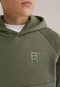 Zöld kapucnis pulóver texturált vállpanelekkel. Hímzett "BLUE RIDGE DENIM" logóval a mellkasán. Puha, pamut keverék anyag.