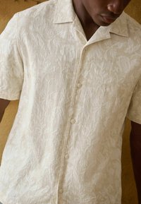 Hombre vistiendo una camisa de botón de manga corta en color crema con una sutil textura jacquard floral, frente a un fondo neutro.