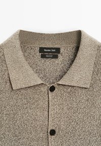Polo en coton beige tricoté avec col et deux boutons noirs, avec une étiquette de marque "Massimo Dutti" au niveau du col.