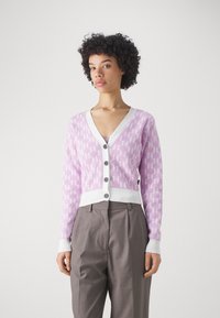 Cárdigan lila claro con un patrón geométrico, escote en V y puños de canalé blancos. Presenta cinco botones negros y un corte corto. Llevado sobre pantalones grises.