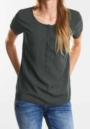 Vrouw draagt een donkergroen kortärmelig blouse met een voorflap, gecombineerd met blauwe spijkerbroek, staand met handen achter haar rug.