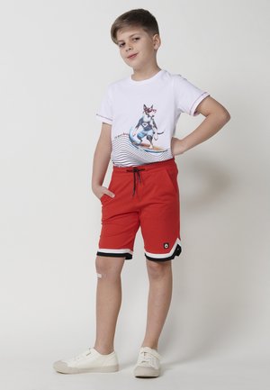 T-shirt bianco con grafica di un cane e onde blu; pantaloni corti rossi con rifiniture nere e bianche, tasche laterali; sneaker bianche.