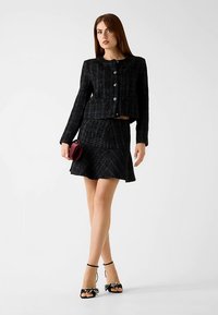 Veste en tweed noire à encolure ronde et boutons, associée à une jupe évasée assortie. Accessoires : un petit sac à main bordeaux et des escarpins noirs.