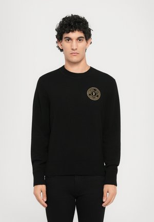 Black knitted sweater with a round neckline and long sleeves, featuring a gold embroidered logo on the left chest and ribbed cuffs and hem.
