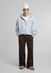 Sweat à capuche gris zippé avec un texte rouge, associé à un pantalon large marron et des chaussures slip-on beiges, sur un fond neutre.