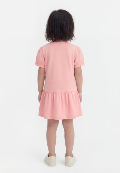 Niña con cabello oscuro hasta los hombros, de espaldas, vistiendo un vestido rosa claro de manga corta y zapatillas blancas sobre un fondo blanco.