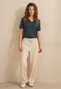 Tiefer blauer Kurzarm-Oberteil mit V-Ausschnitt, kombiniert mit cremefarbenen, elastischen weit geschnittenen Hosen. Das Model trägt weiße Sneaker.