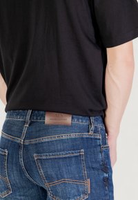 Persona que lleva vaqueros oscuros con costuras naranjas y una camiseta negra, mostrando la parte trasera y la etiqueta de cuero de Armani Exchange en la cinturilla.